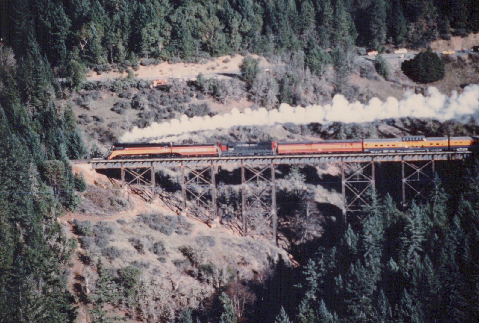A steam locomotive pulling passenger cars crosses a high trestle bridge through a forested landscape with a plume of steam rising from its stack.