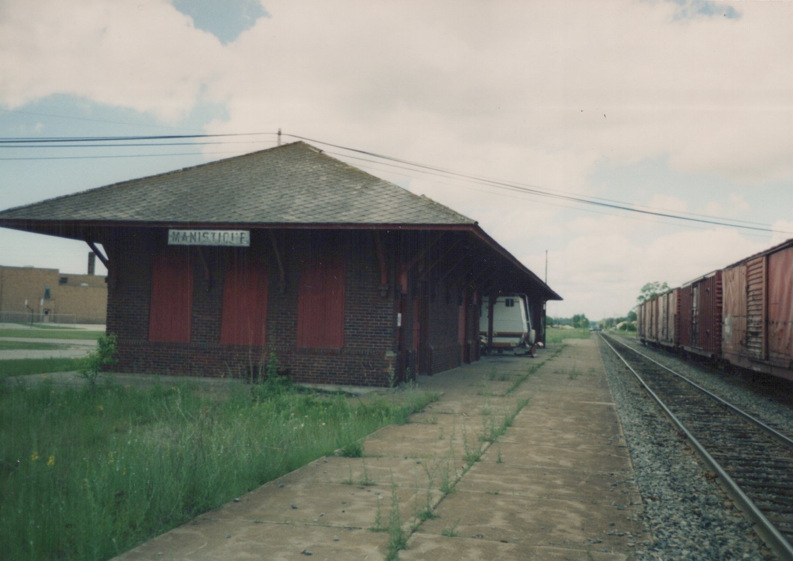 A brick train station named Manistique with overgrown weeds on the platform, a freight train to the right, and a white trailer parked under the roof.