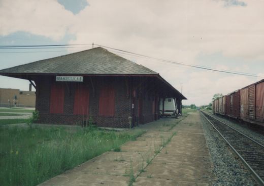 A brick train station named Manistique with overgrown weeds on the platform, a freight train to the right, and a white trailer parked under the roof.