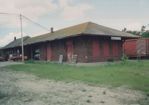 A brick train station with a sign reading 'MANISTIQUE', a red freight car parked beside it, and several vehicles parked under the station's overhang on a cloudy summer day.