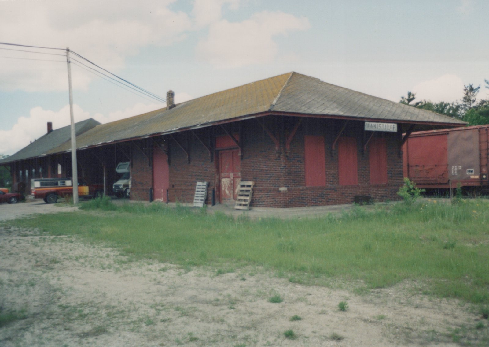 A brick train station with a sign reading 'MANISTIQUE', a red freight car parked beside it, and several vehicles parked under the station's overhang on a cloudy summer day.