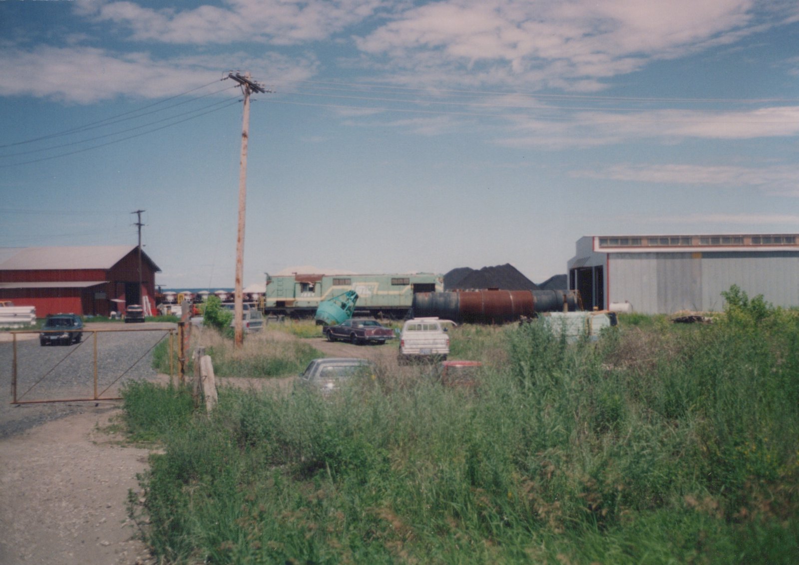 A vintage photo showing a green and yellow diesel locomotive parked in an industrial yard with overgrown grass, a red barn, and several cars in the 1970s.