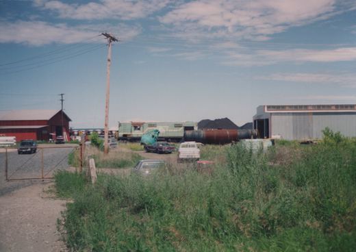 A vintage photo showing a green and yellow diesel locomotive parked in an industrial yard with overgrown grass, a red barn, and several cars in the 1970s.