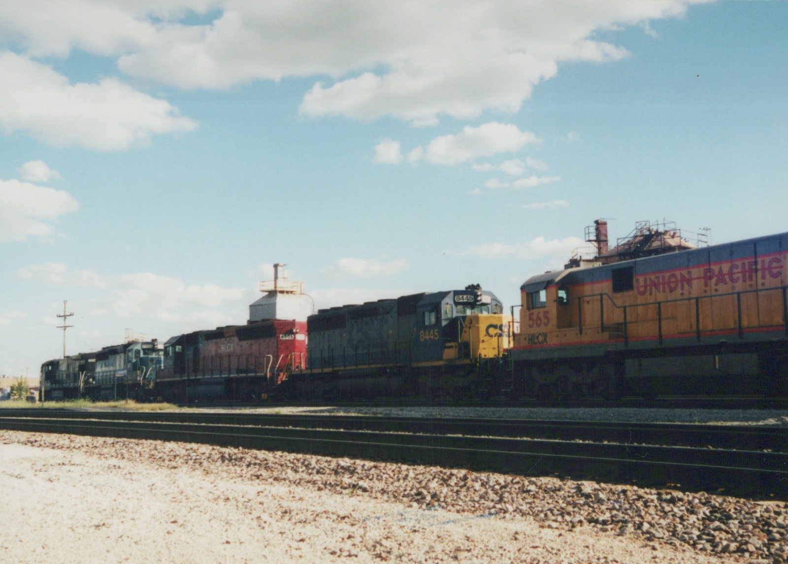 A side view of several diesel locomotives from different operators, including CSX, Union Pacific, and HLCK, lined up on parallel tracks in a rail yard under a partly cloudy sky.