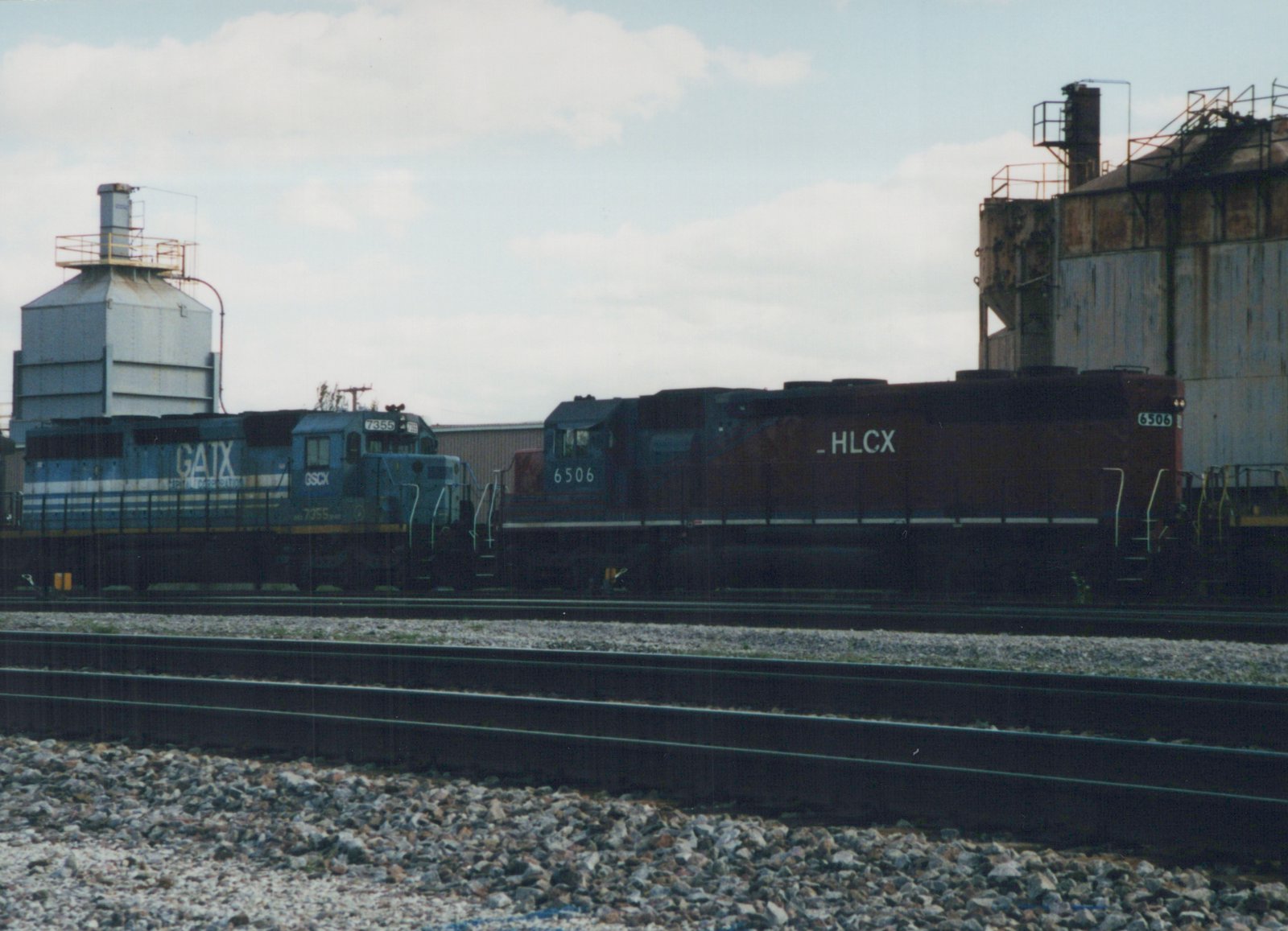 Two diesel locomotives, a blue GATX GP38-2 numbered 7355 and a dark HLCX GP9 numbered 6506, are parked next to each other on parallel tracks in a rail yard under an overcast sky.