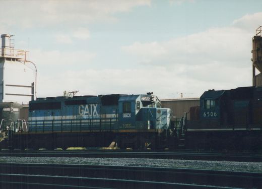 Side view of a blue and white GATX SD40-2 locomotive, number 7355, parked on tracks in a rail yard with other locomotives and industrial buildings in the background.