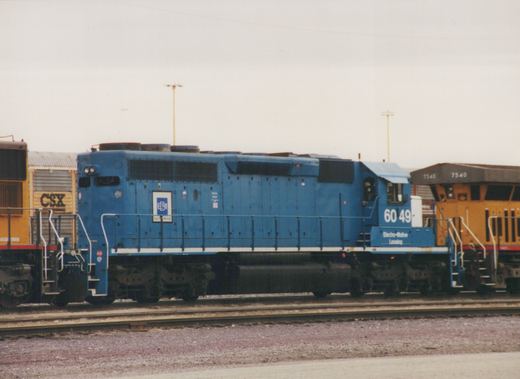 A blue diesel locomotive with the number 6049 and lettering 'Electro-Motive Leasing' is parked in a rail yard, with a yellow CSX locomotive nearby.