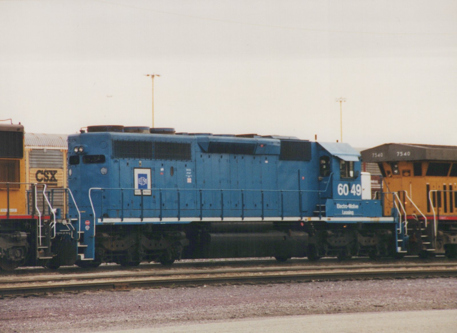 A blue diesel locomotive with the number 6049 and lettering 'Electro-Motive Leasing' is parked in a rail yard, with a yellow CSX locomotive nearby.