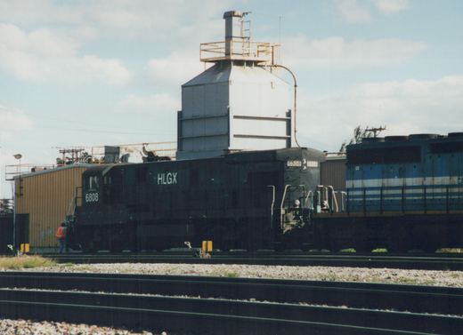 A black HLGX 6808 diesel locomotive on tracks in a rail yard, with a person standing near its steps and a blue locomotive partially visible behind it.