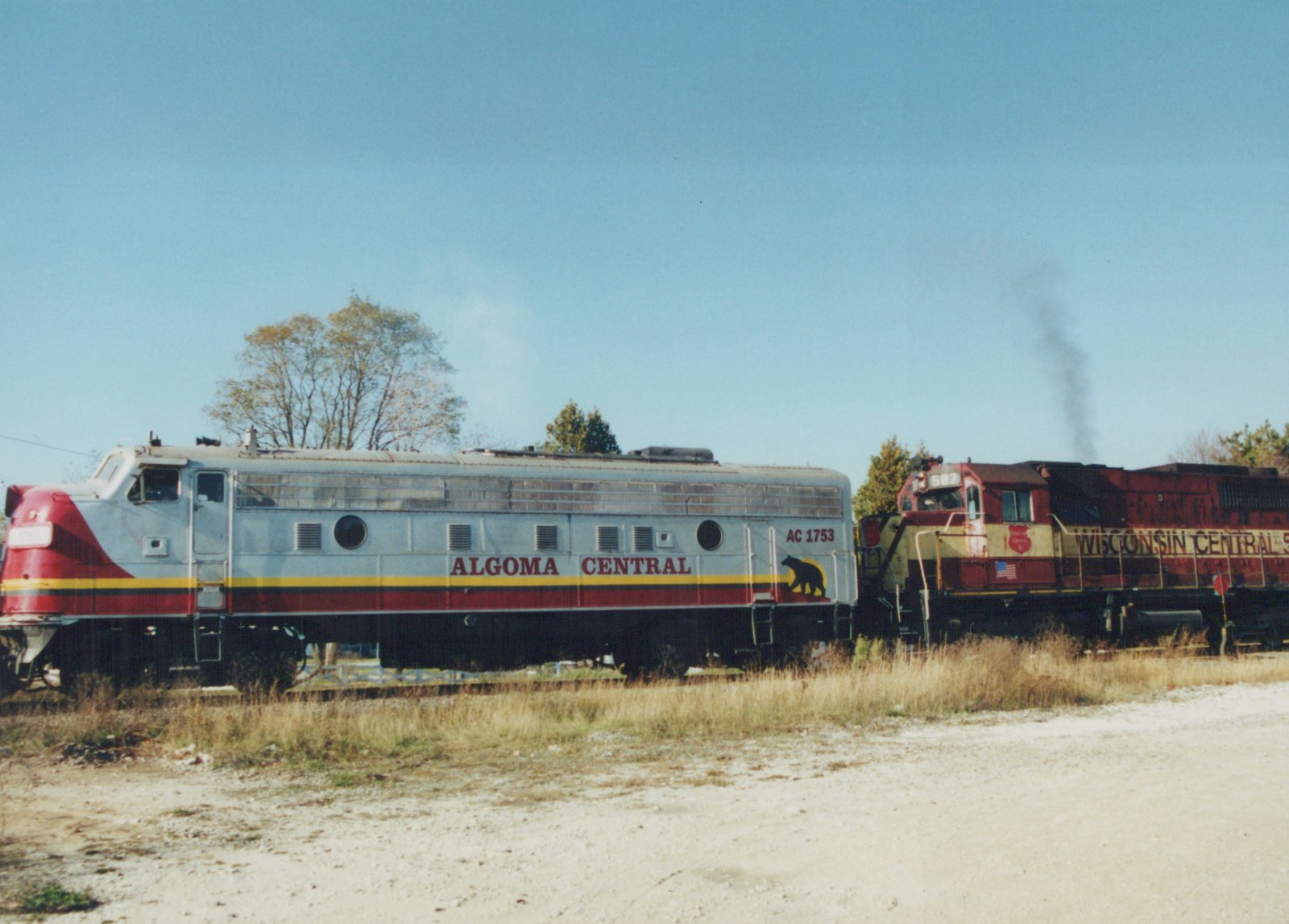 Side view of an Algoma Central self-propelled railcar numbered AC 1753 next to a Wisconsin Central locomotive numbered 587, on a private right-of-way with autumn trees in the background under a clear sky.