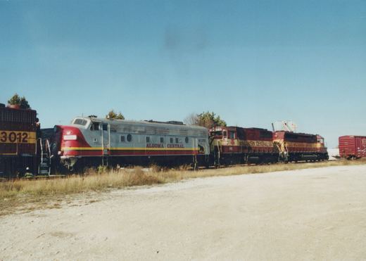 A silver Algoma Central self-propelled railcar and two maroon Wisconsin Central diesel locomotives sit on parallel tracks in a yard with dry grass.