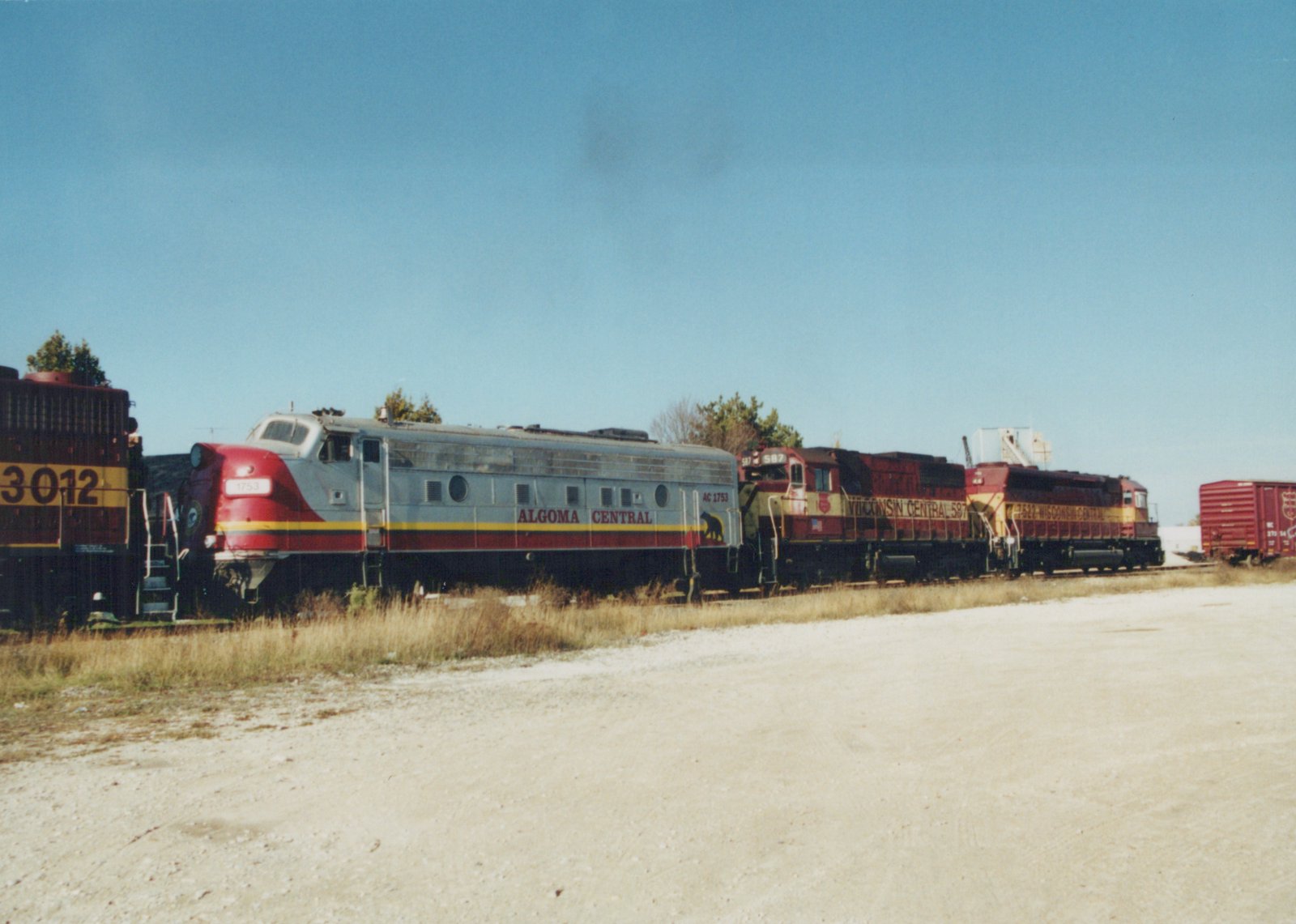 A silver Algoma Central self-propelled railcar and two maroon Wisconsin Central diesel locomotives sit on parallel tracks in a yard with dry grass.