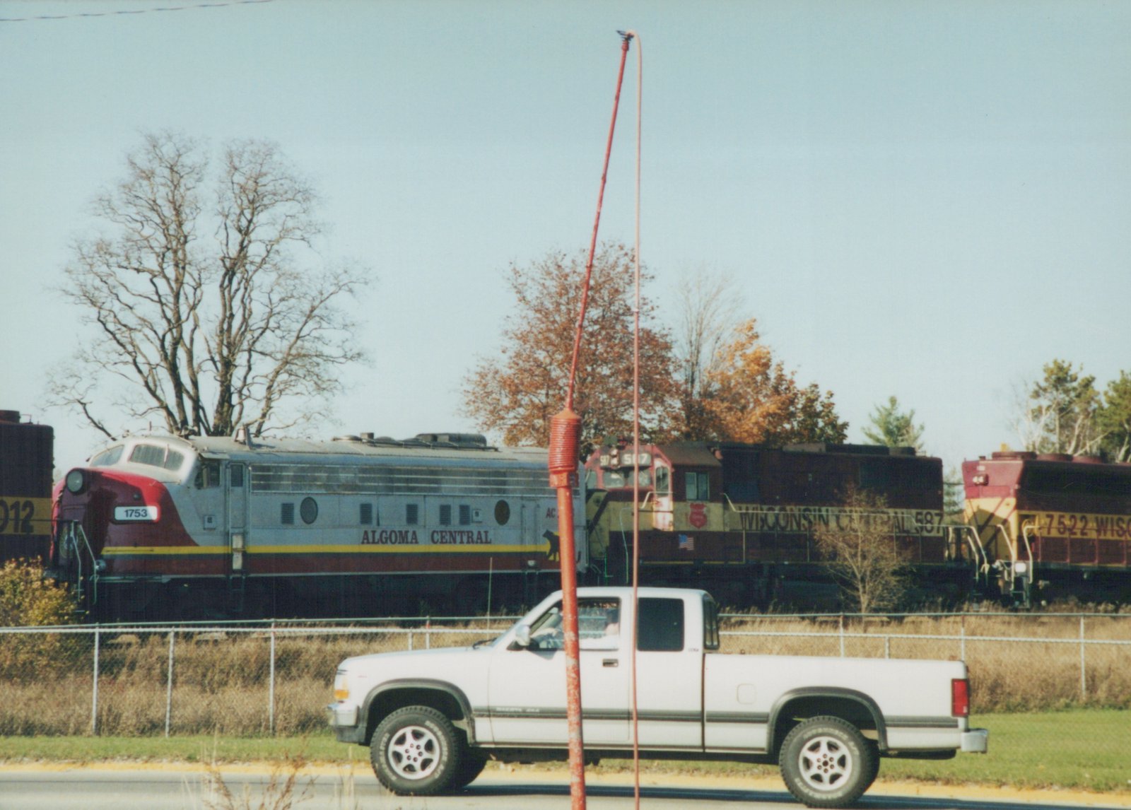 Side view of a train with an Algoma Central locomotive and two Wisconsin Central locomotives at a grade crossing, with a white pickup truck on the road in front and autumn trees in the background.