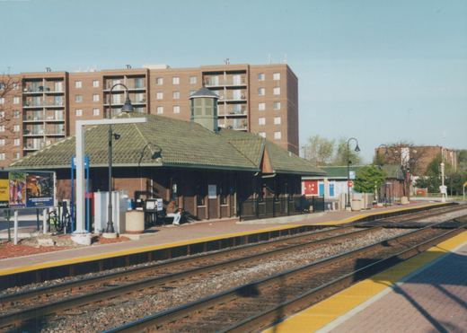 A sunny day at a railroad station with a covered platform, two people waiting on a bench, and apartment buildings visible behind the station.