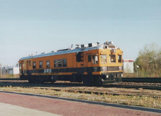 A yellow and black railcar labeled SRS 137 sits on railroad tracks next to a brick sidewalk, with trees and a clear sky in the background.