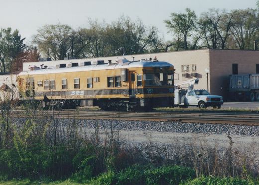 A yellow and black self-propelled railcar, number 137 for Speary Rail Service, is parked on tracks beside a building and a white utility truck.
