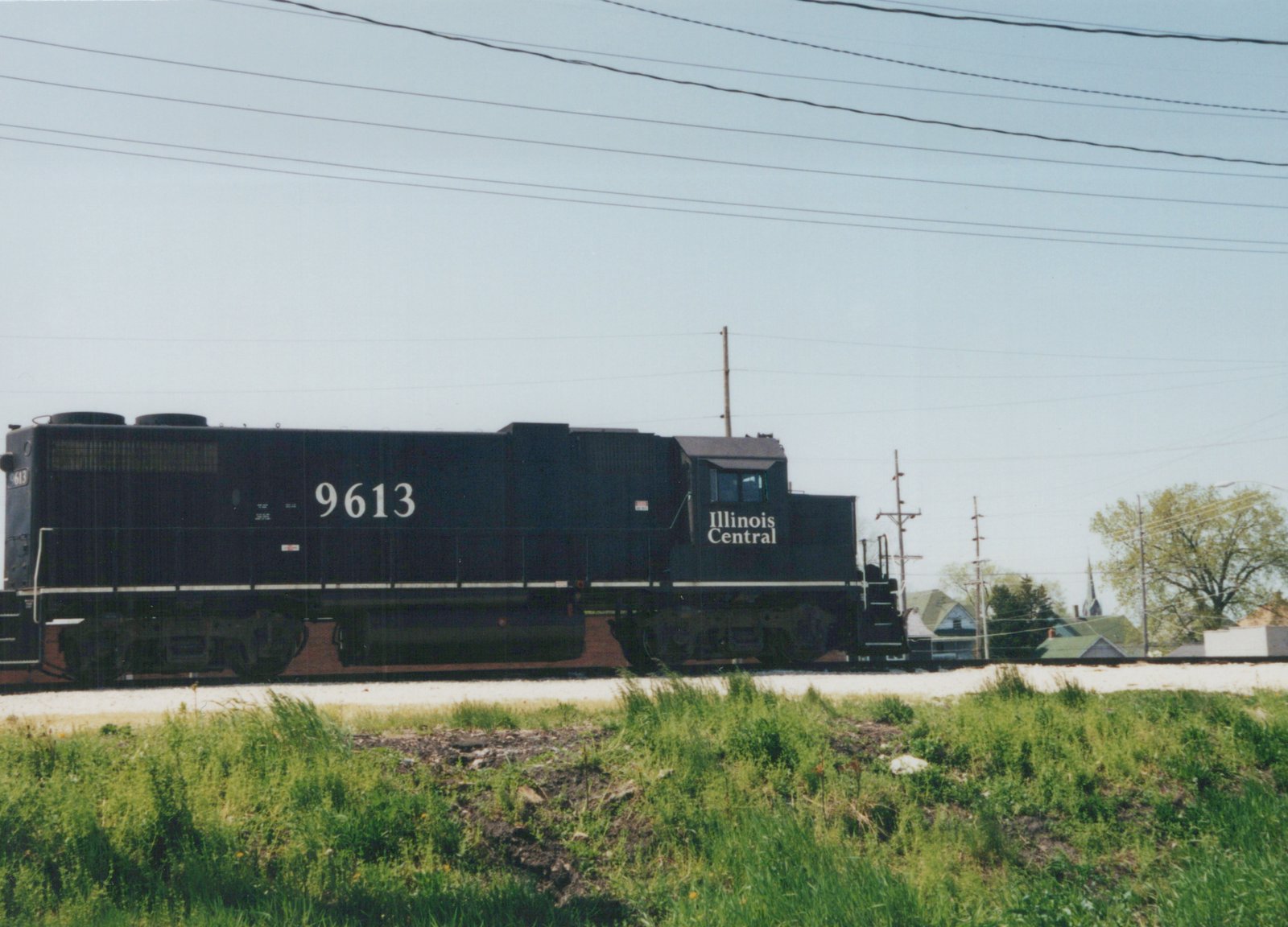 Side view of a black Illinois Central diesel locomotive numbered 9613 on a grassy track with houses and power lines in the background.