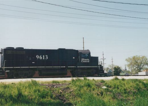 Side view of a black Illinois Central diesel locomotive numbered 9613 on a grassy track with houses and power lines in the background.