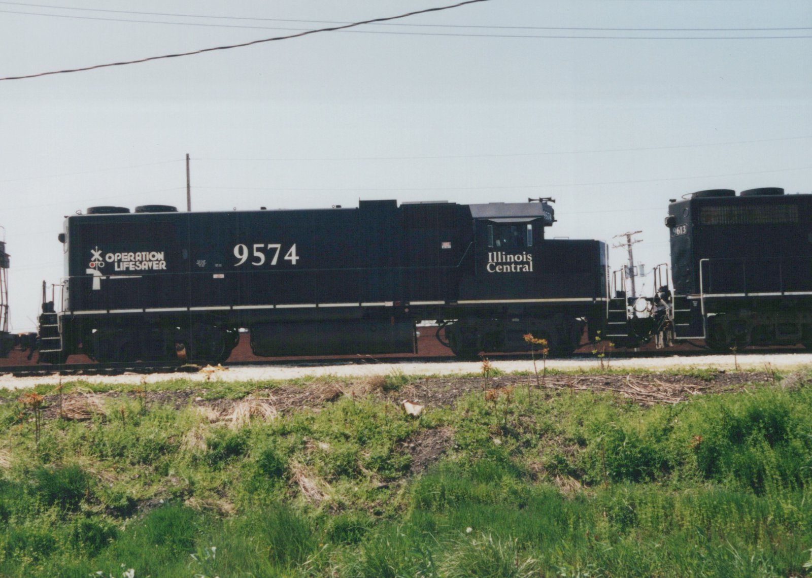 Side view of a black Illinois Central diesel locomotive with 'Operation Lifesaver' lettering, pulling another locomotive on a track beside green grass under a clear sky.