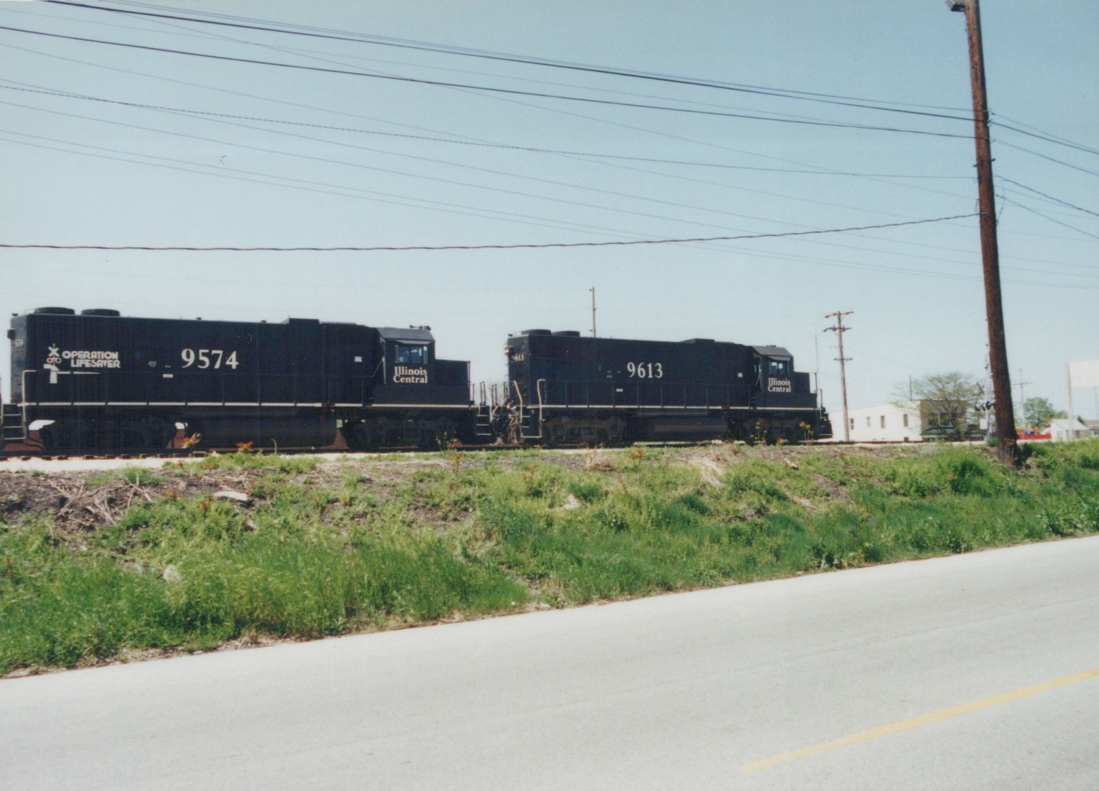 Two black Illinois Central locomotives, numbered 9574 and 9613, stand on railroad tracks next to a grassy embankment and a paved road. The lead locomotive displays an Operation Lifesaver logo.