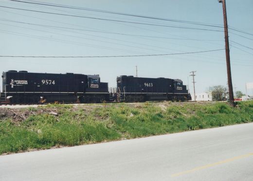 Two black Illinois Central locomotives, numbered 9574 and 9613, stand on railroad tracks next to a grassy embankment and a paved road. The lead locomotive displays an Operation Lifesaver logo.