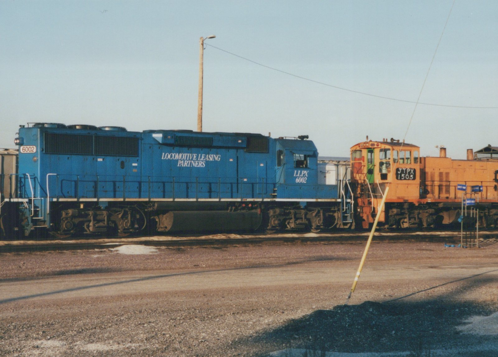 Two diesel locomotives, one blue and one orange, are parked side by side in a gravel yard under a clear sky. The blue locomotive displays the number 6002 and the text 'LOCOMOTIVE LEASING PARTNERS'. The orange locomotive displays the number 1503 and the reporting mark JTPX.
