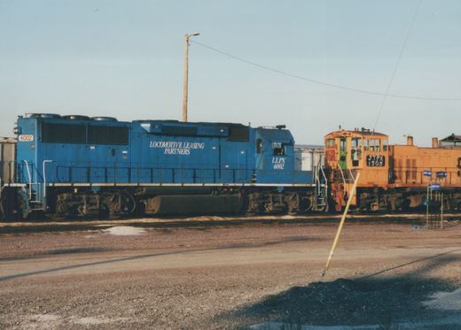 Two diesel locomotives, one blue and one orange, are parked side by side in a gravel yard under a clear sky. The blue locomotive displays the number 6002 and the text 'LOCOMOTIVE LEASING PARTNERS'. The orange locomotive displays the number 1503 and the reporting mark JTPX.