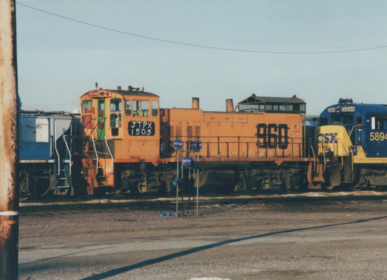 Two diesel locomotives, one orange with JTPX 1503 and the other blue with CSX 5894, are parked next to each other in a gravel rail yard under a clear sky.
