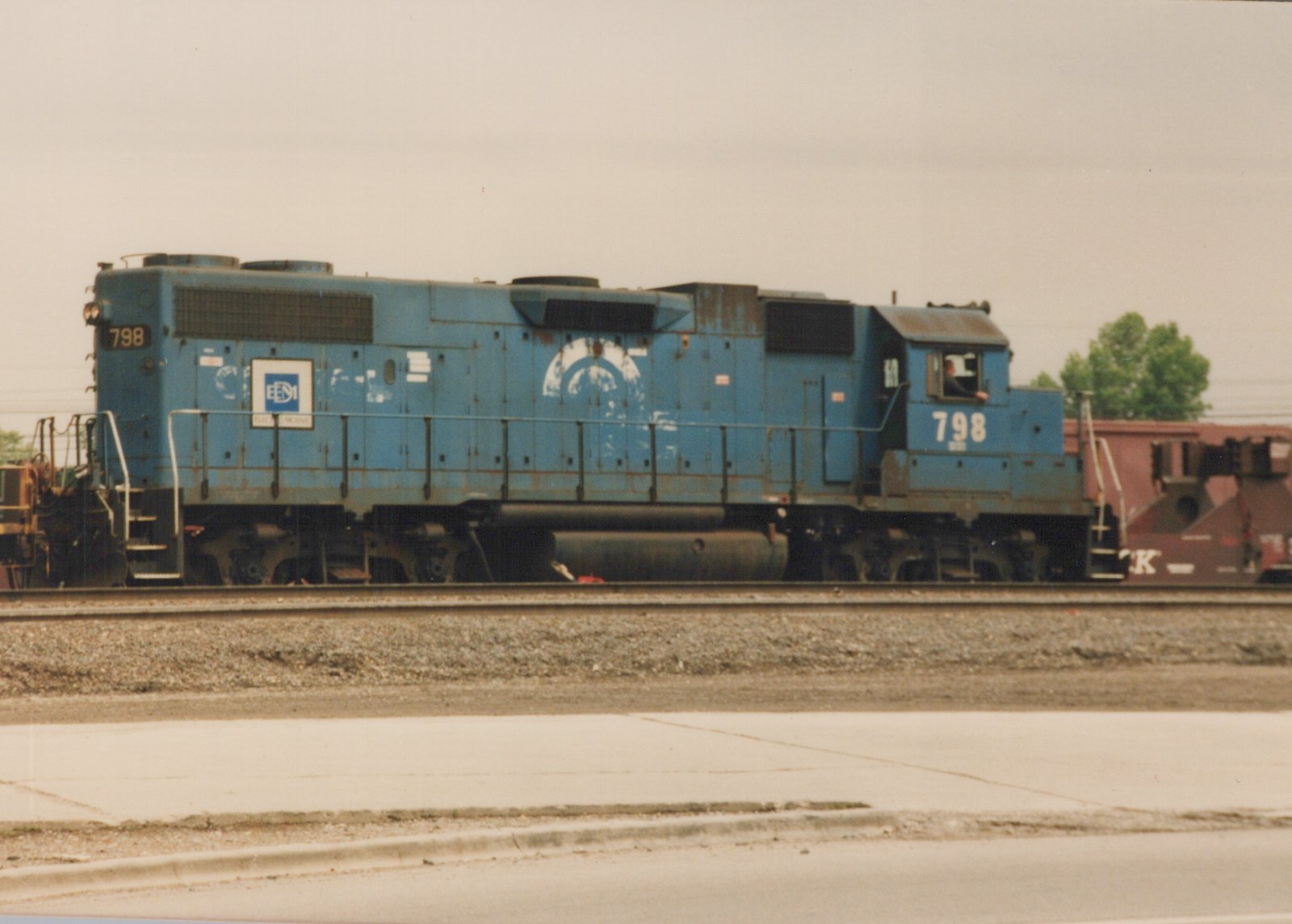 A blue diesel locomotive with the number 798 and E&M lettering is parked on railroad tracks in a yard with a freight car behind it.