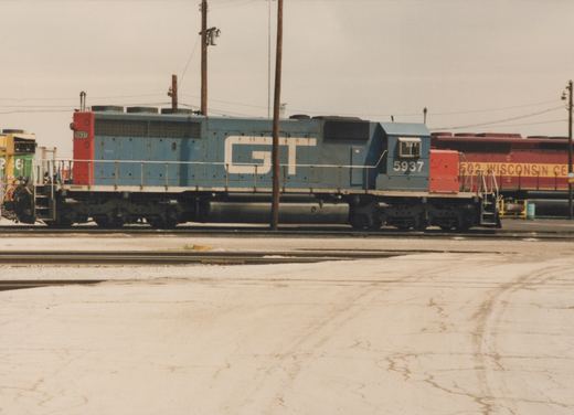 A side view of a blue and red GT diesel locomotive numbered 5937, parked in a rail yard with another locomotive in the background.