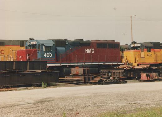 Two diesel locomotives, one red and blue and one yellow, are parked side by side in a gravel rail yard under an overcast sky.