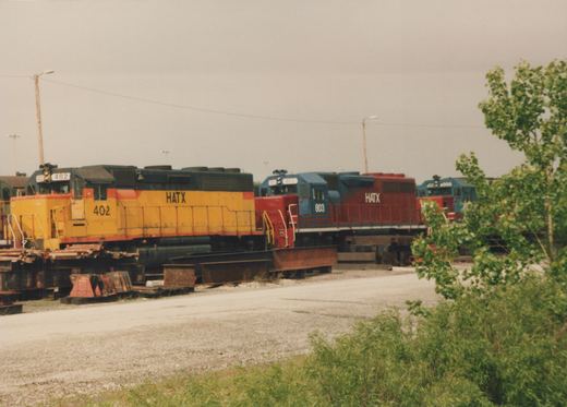 Three diesel locomotives with HATX lettering, numbered 402, 803, and 4000, are parked in a gravel yard, surrounded by green vegetation and utility poles.
