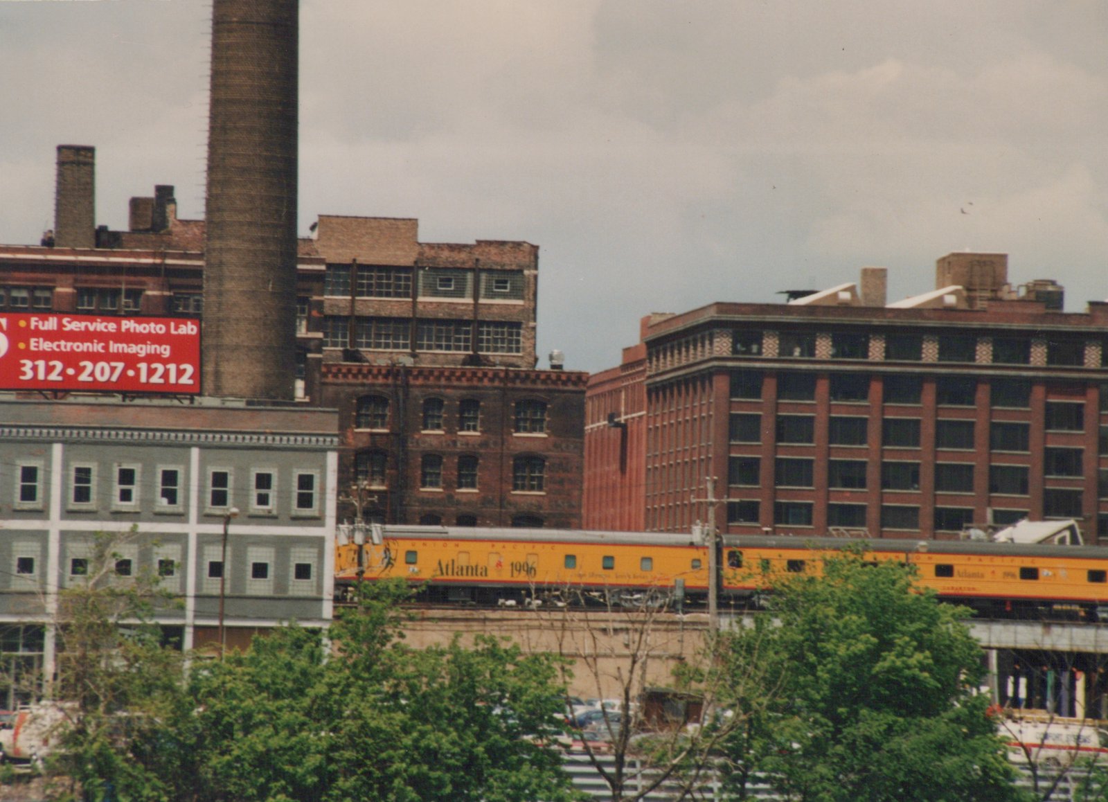 Two yellow Union Pacific passenger cars with 'Atlanta 1996' graphics are seen on an elevated track, with industrial brick buildings and a red billboard in the background under an overcast sky.