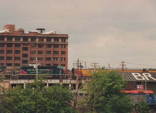 A green Union Pacific locomotive with '1996 Olympic Torch Relay' graphics moves across an elevated track, with a red brick building and trees in the foreground.