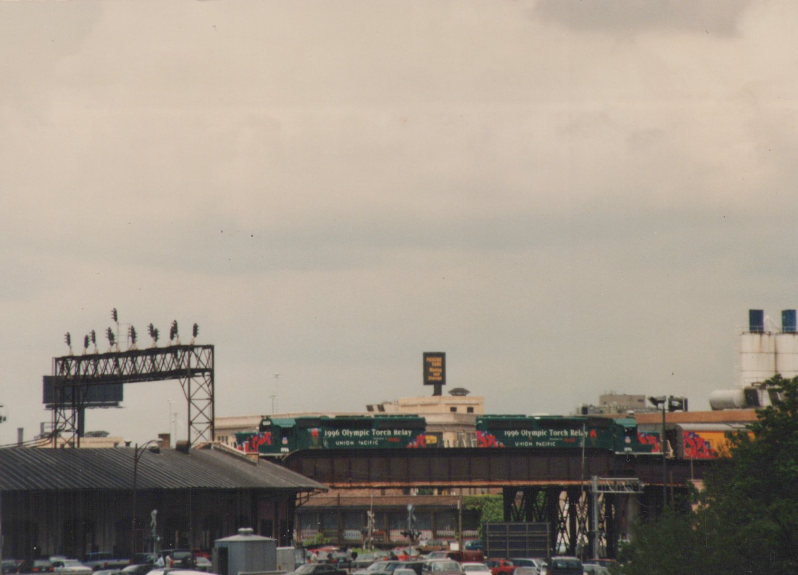 A green Union Pacific locomotive with '1996 Olympic Torch Relay' lettering travels on an elevated track over a busy city street with cars and buildings below.