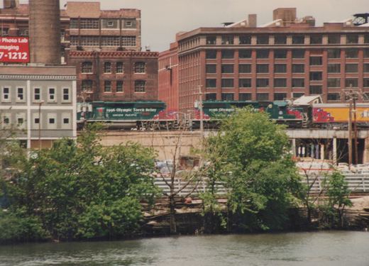 A green Union Pacific locomotive with '1996 Olympic Torch Relay' lettering travels on an elevated track above a river, with brick buildings in the background.
