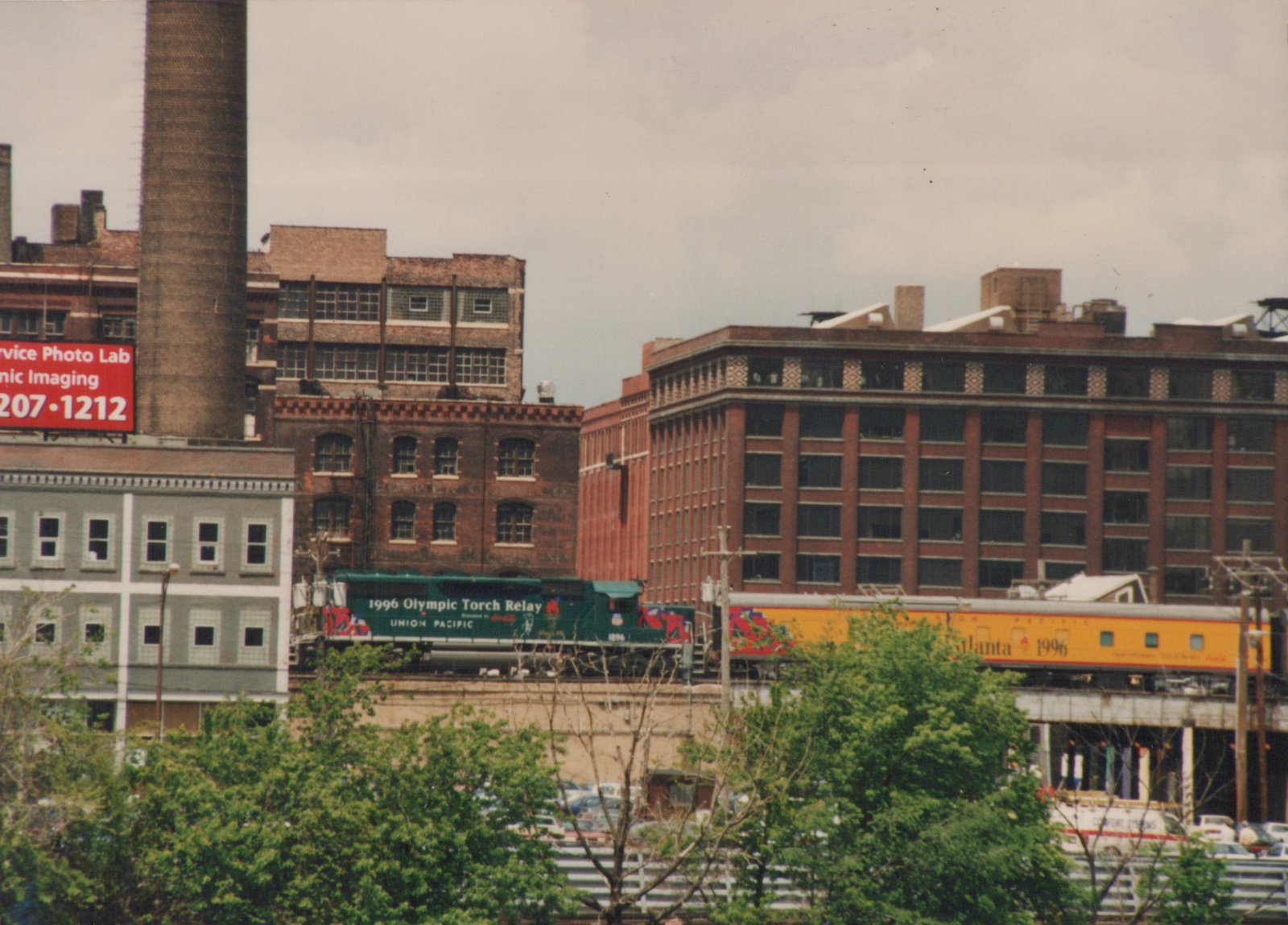 A green and yellow Union Pacific train for the 1996 Olympic Torch Relay travels on an elevated track through a city with brick buildings and green trees.