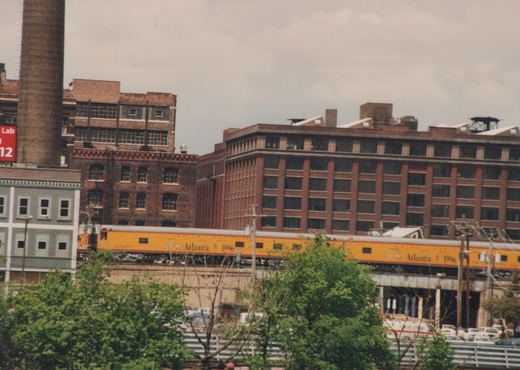 Two yellow Union Pacific passenger cars with 'Atlanta 1996' lettering are on an elevated concrete track, with large brick buildings and green trees in the background under an overcast sky.