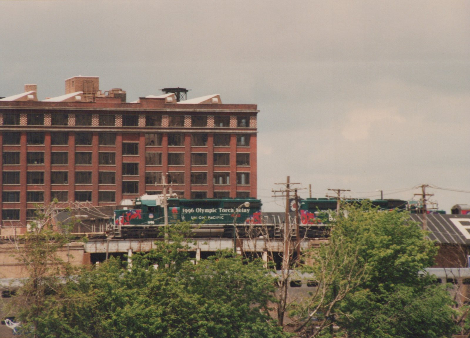 A green Union Pacific locomotive with '1996 Olympic Torch Relay' lettering is on an elevated track against a backdrop of a red brick building and an overcast sky.