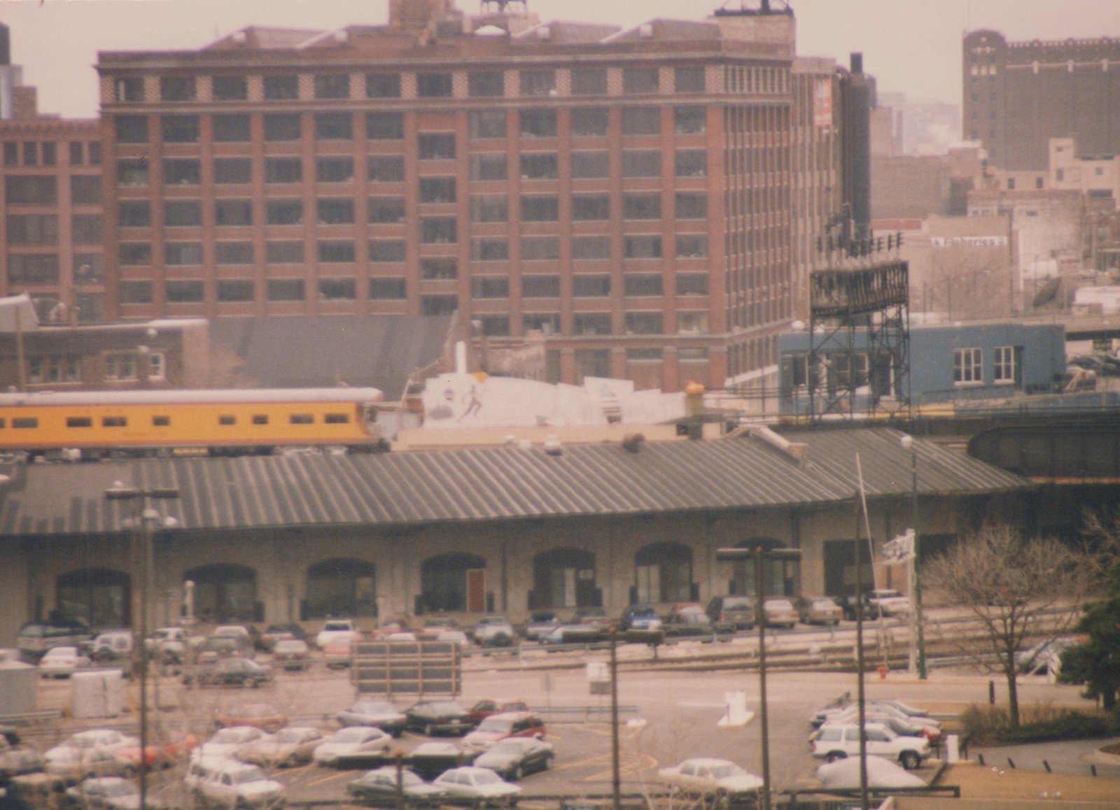 A long, yellow railroad passenger car moves along an elevated track above a building with arched openings and a large parking lot filled with cars, with a tall brick building in the background.