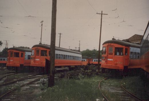 A row of three orange and grey interurban cars, numbers 452, 455, and 456, belonging to the Chicago Aurora and Elgin Railroad, sit on tracks in a grassy yard under a cloudy sky.
