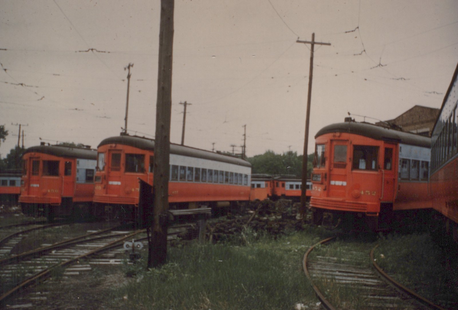 A row of three orange and grey interurban cars, numbers 452, 455, and 456, belonging to the Chicago Aurora and Elgin Railroad, sit on tracks in a grassy yard under a cloudy sky.