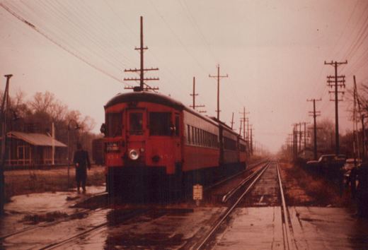 A red and cream CA&E interurban car numbered 171 is stopped at a rural station platform with overhead wires, a person walking nearby, and a car parked in the background.