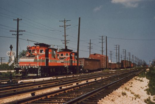 Two bright orange CA&E electric locomotives lead a freight train on multiple tracks under overhead wires on a sunny day.