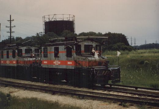 Two vintage Chicago Aurora and Elgin interurban railcars, 3003 and 3004, travel on a track bordered by grass and trees, with a large water tank visible behind them under an overcast sky.