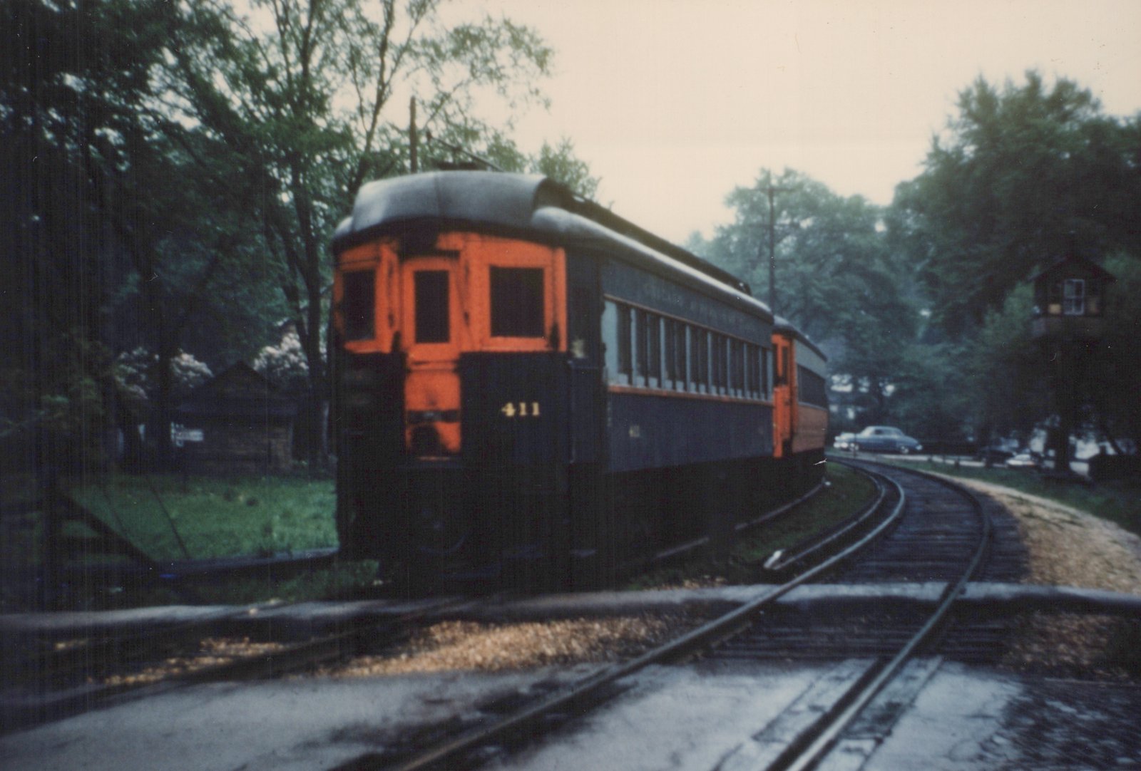 An orange and dark blue interurban car, number 411, with 'Chicago & North Western' lettering, is rounding a curve on a private right-of-way with overhead wires, passing a wooden signal tower and a parked automobile in the background.