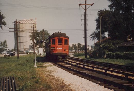 A red electric interurban train car numbered 34 is on a set of tracks next to a grassy area, with a water tower and vintage cars in the background.