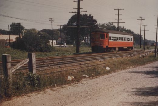 An orange and white interurban railcar on tracks beside a gravel road, with trees and utility poles in the background.