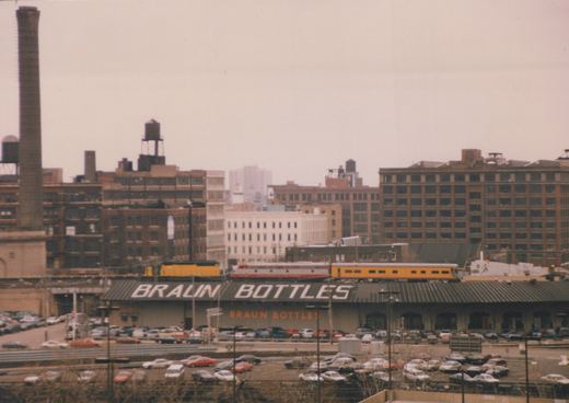 An elevated train with a silver and red locomotive and a yellow passenger car travels over a street with a large 'Braun Bottles' sign on a building and parked cars below, under an overcast sky.