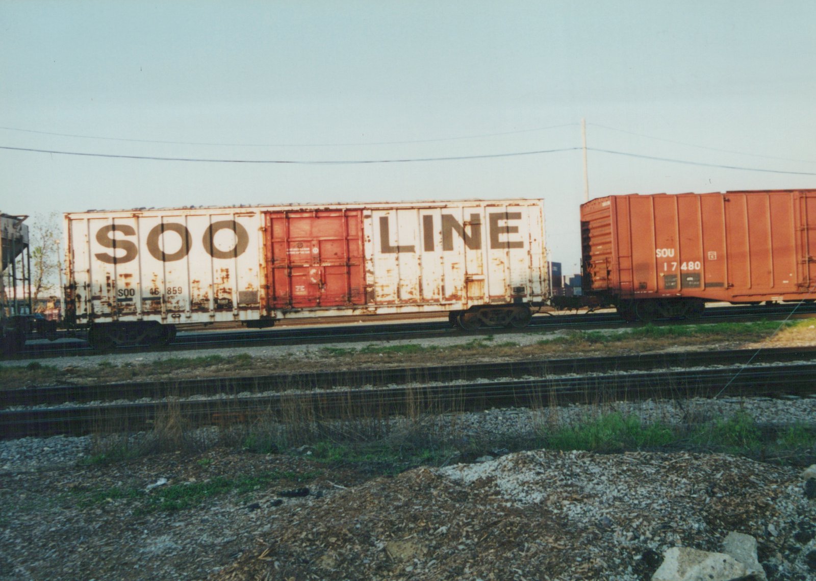 Two freight cars, one white with 'SOO LINE' lettering and the other orange-red with 'SOU' markings, are parked on adjacent tracks in a gravel yard.